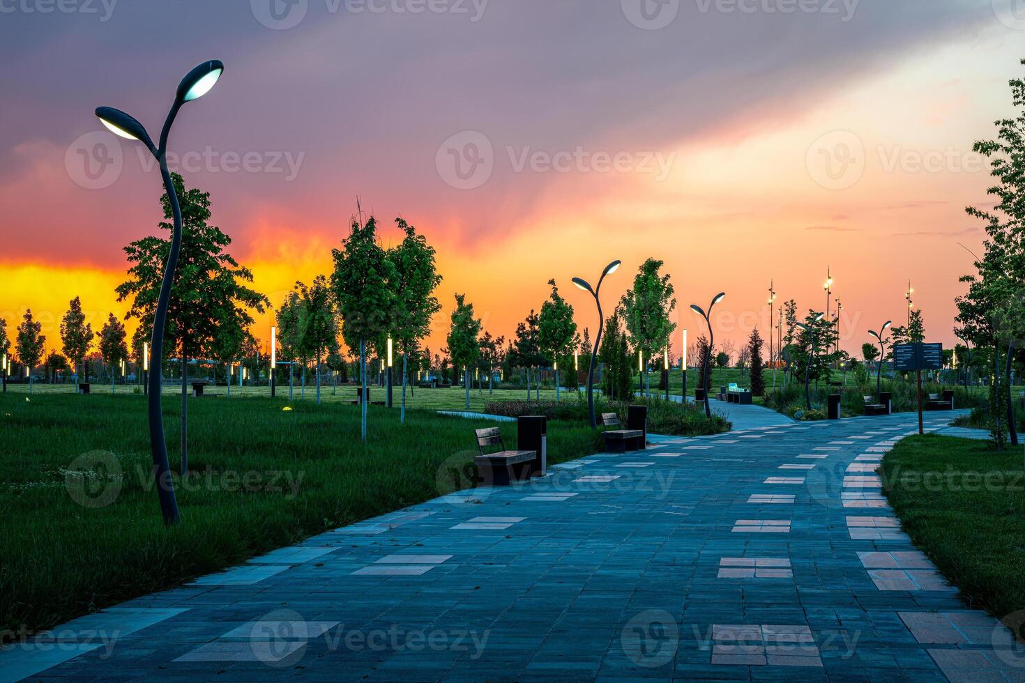 Stadt Park im früh Sommer- oder Frühling mit Gehweg, Laternen, jung Grün Rasen, Bäume und dramatisch wolkig Himmel auf ein Sonnenuntergang oder Sonnenaufgang. foto