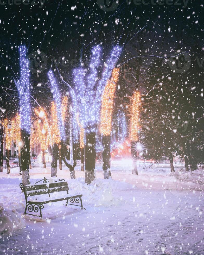 Schneefall im ein Winter Park beim Nacht mit Dekorationen, glühend Laternen, Pflaster bedeckt mit Schnee und Bäume. Jahrgang Film ästhetisch. foto