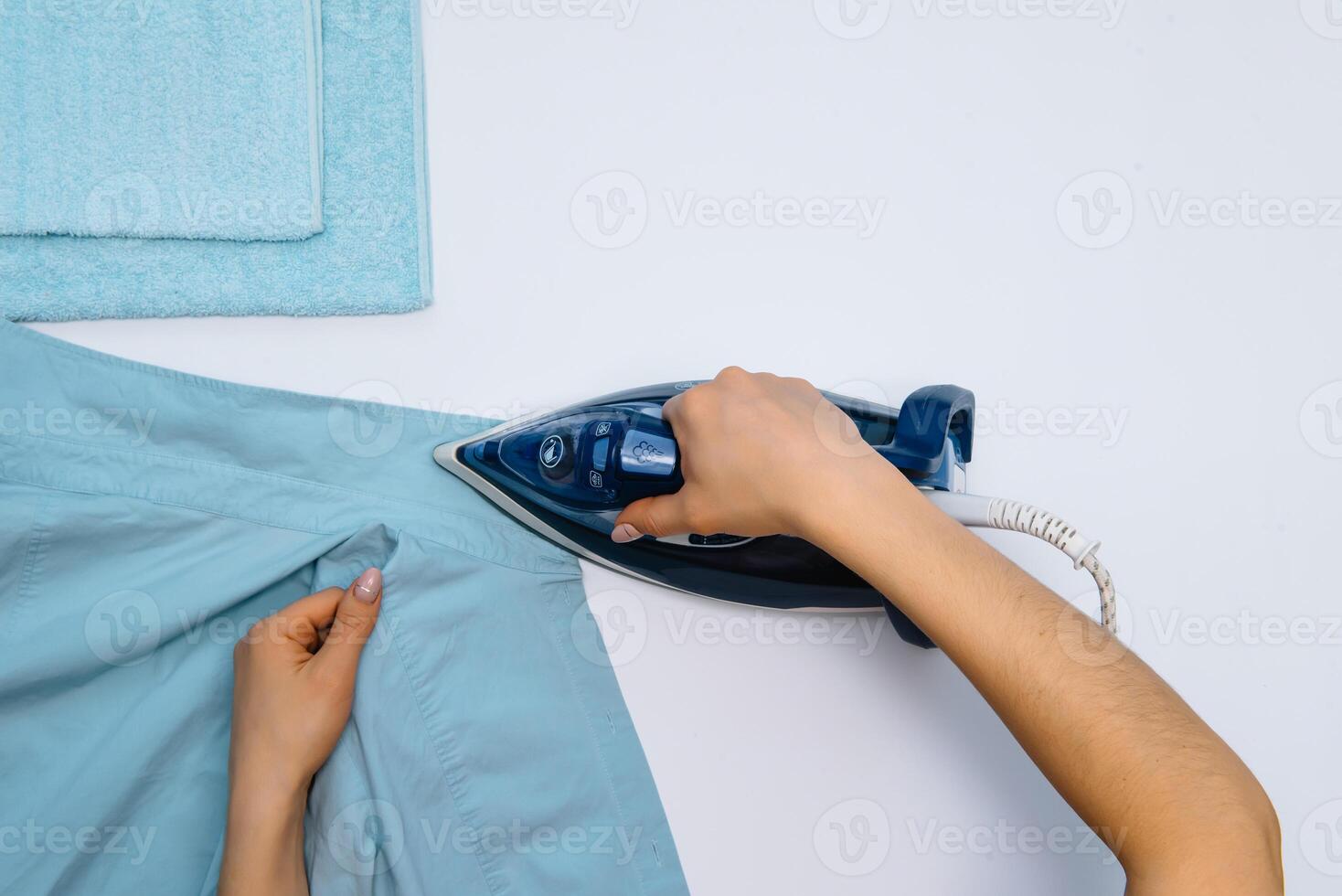 weiblich Hand Bügeln Kleider oben Aussicht isoliert auf Weiß Hintergrund. jung Frau mit Eisen Bügeln des Mannes Hemd gesehen von über während Hausarbeit. Blau Eisen auf Weiß Tabelle foto