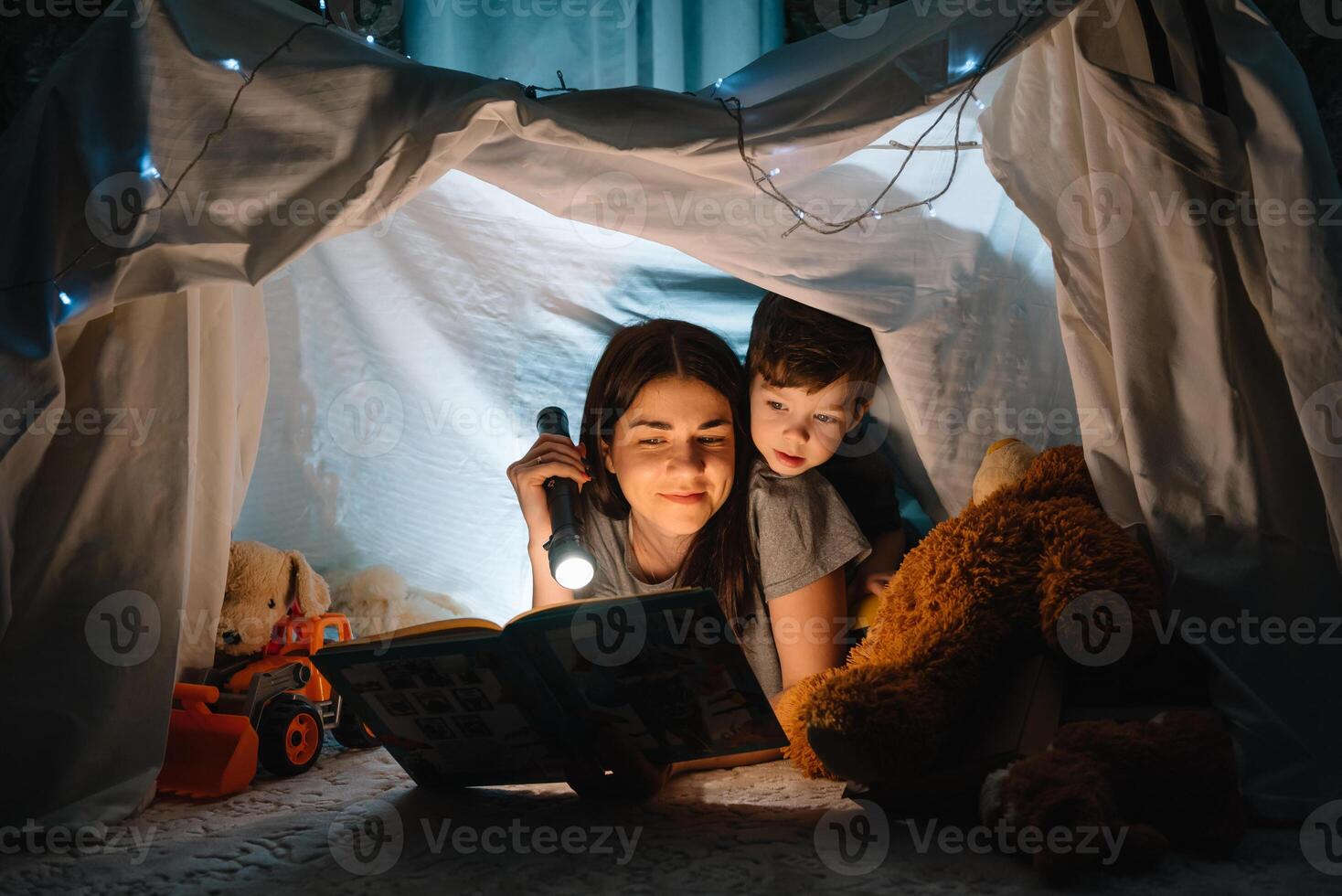 glücklich Familie Mutter und Kind Sohn lesen ein Buch mit ein Taschenlampe im ein Zelt beim heim. Familie Konzept. foto