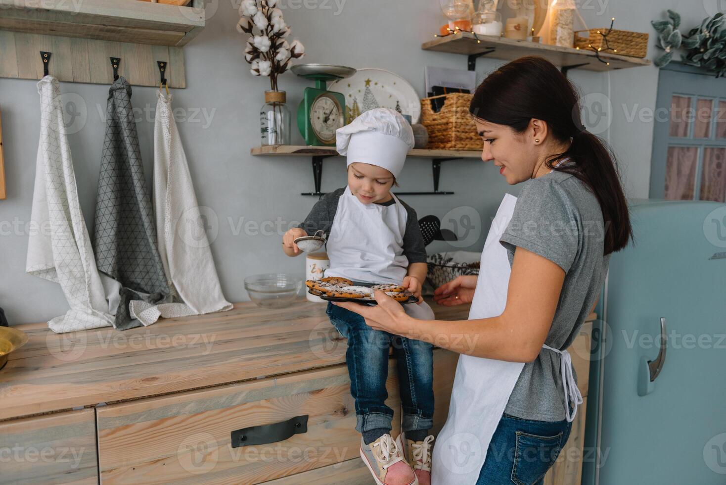 jung glücklich Mama und ihr Baby Koch Kekse beim Zuhause im das Küche. Weihnachten hausgemacht Lebkuchen. süß Junge mit Mutter im Weiß Uniform und Hut gekocht Schokolade Kekse foto