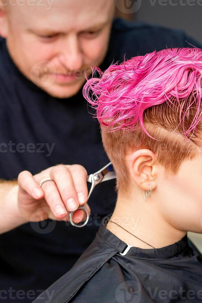 Frau haben ein Neu Haarschnitt. ein männlich Friseur ist Schneiden gefärbt Rosa kurz Haar mit Schere im ein Haar Salon. foto