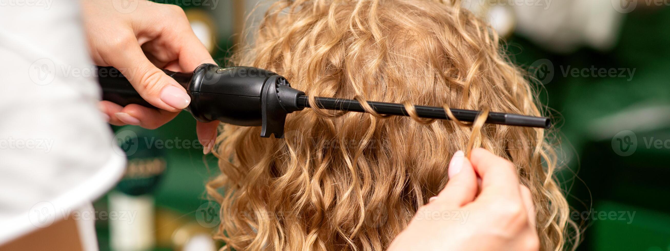 Hände von Friseur locken wellig Haar von jung Frau mit ein Eisstockschießen Eisen zum Haar Locken im das Schönheit Salon Rückseite Sicht. foto