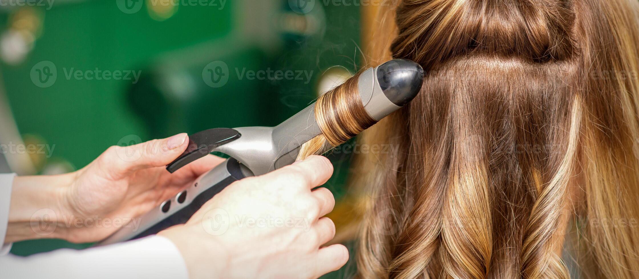 Erstellen Locken mit Eisstockschießen Bügeleisen. Friseur macht ein Frisur zum ein jung Frau mit lange rot Haar im ein Schönheit Salon. foto