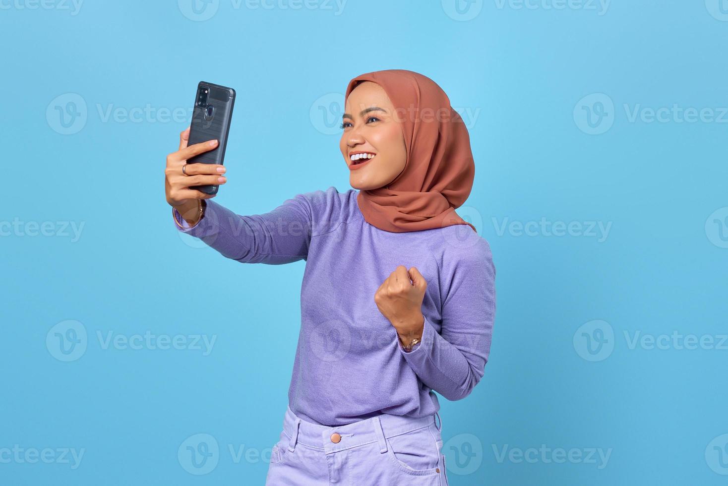fröhliche junge asiatische frau, die ein handy benutzt, während sie den sieg auf blauem hintergrund feiert foto