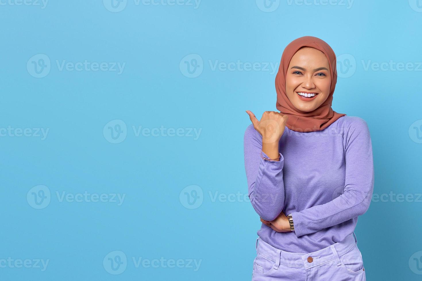 Porträt einer lächelnden jungen asiatischen Frau, die mit dem Daumen auf den Kopierraum auf blauem Hintergrund zeigt foto