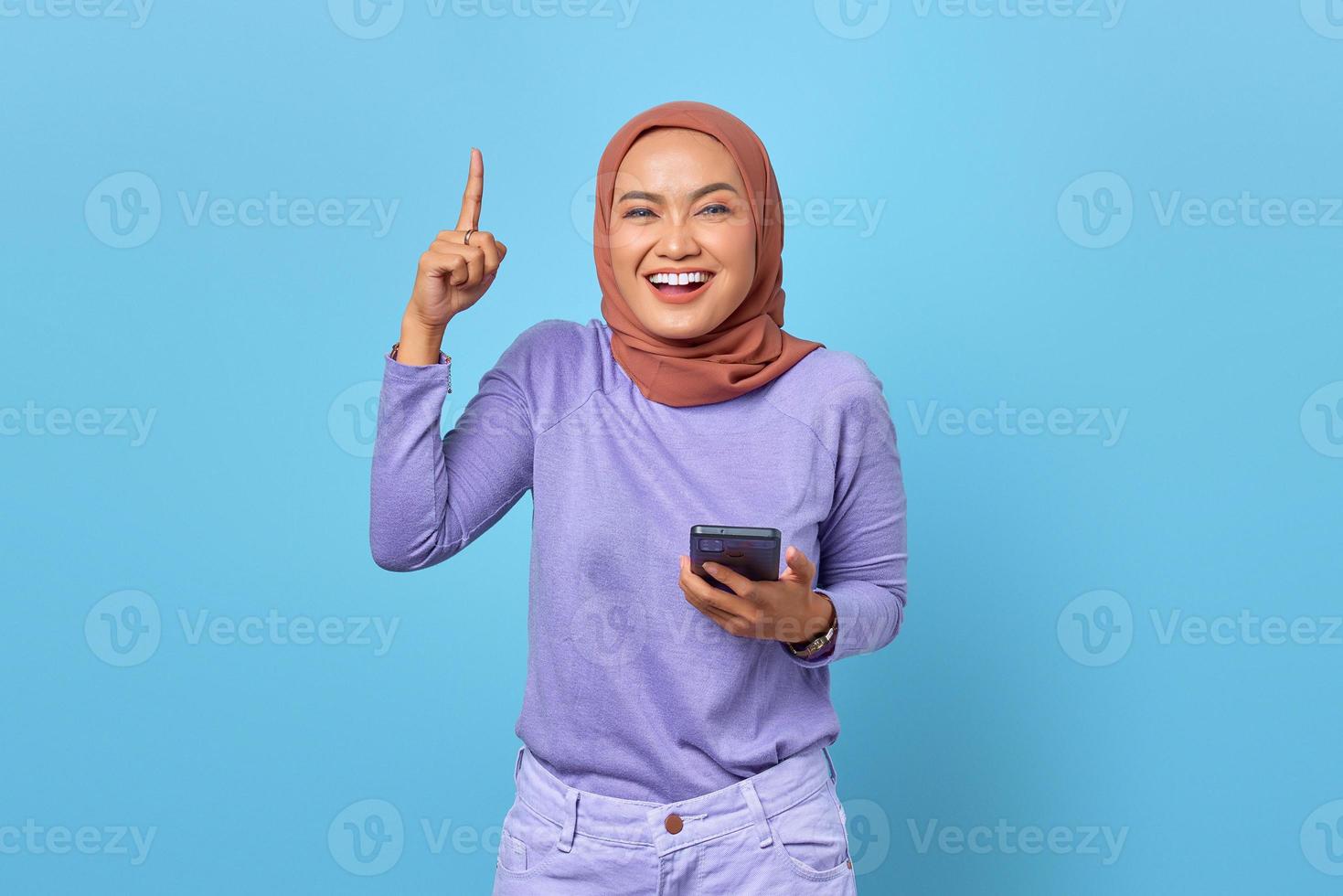 Porträt einer lächelnden jungen asiatischen Frau, die Handy hält, während der Finger auf blauem Hintergrund erhoben wird foto