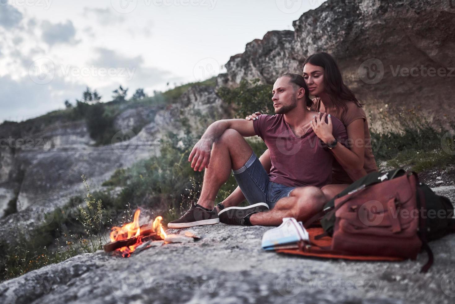 umarmendes Paar mit Rucksack, das in der Nähe des Feuers auf dem Berg sitzt und die Aussicht auf die Küste eines Flusses oder Sees genießt. Reisen entlang von Bergen und Küste, Freiheit und aktives Lifestyle-Konzept foto