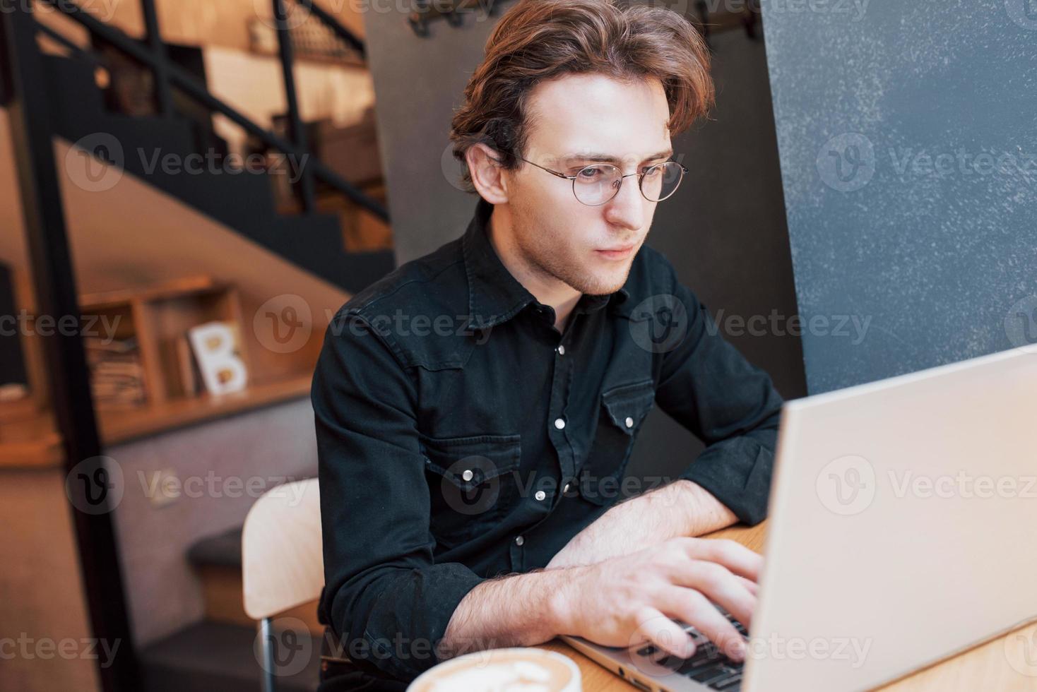 Kreativer Mann-Designer, der an seinem Laptop arbeitet, während er auf Bestellungen in seinem Lieblingscafé im Haus wartet, männlicher Student, der beim morgendlichen Frühstück im modernen Café-Interieur am Net-Book arbeitet foto