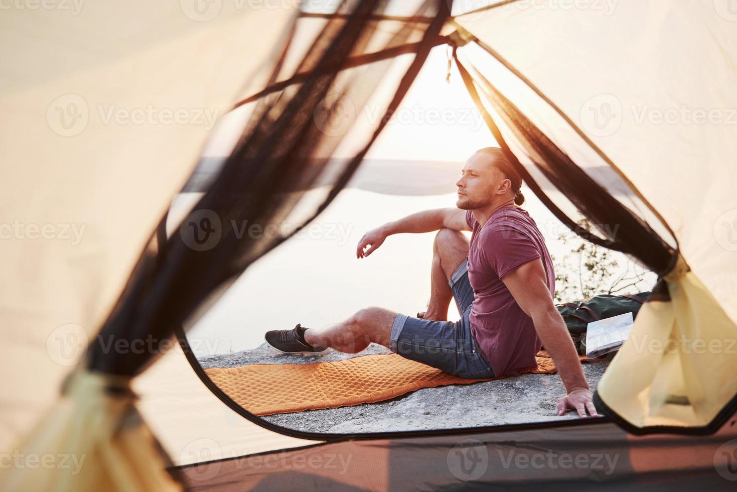 Blick vom Zelt des Reisenden mit Rucksack, der auf dem Berg sitzt und die Aussicht auf die Küste eines Flusses oder Sees genießt. Reisen entlang von Bergen und Küste, Freiheit und aktives Lifestyle-Konzept foto