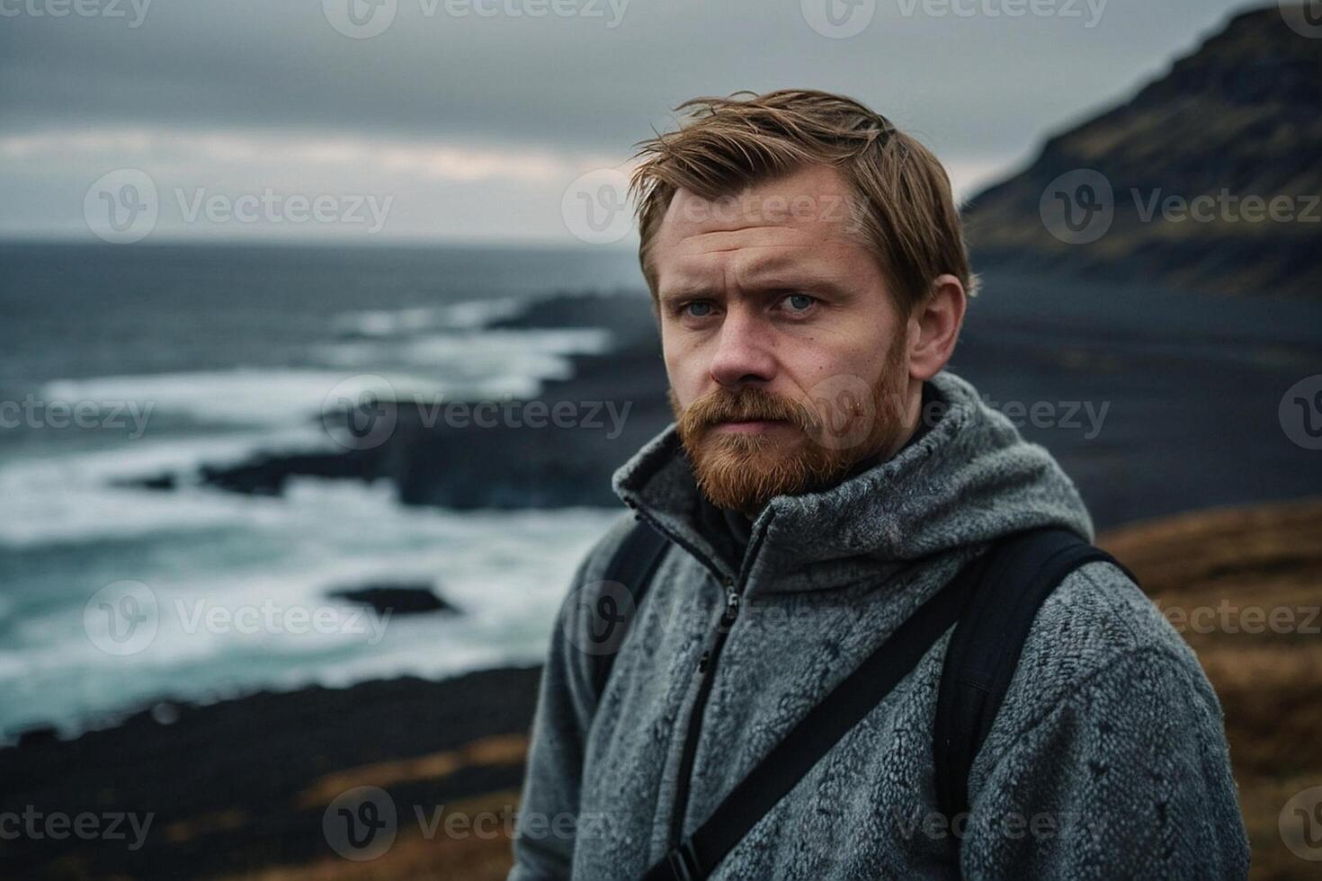 ein Mann mit ein Bart und ein grau Jacke Stehen auf ein Cliff mit Blick auf das Ozean foto