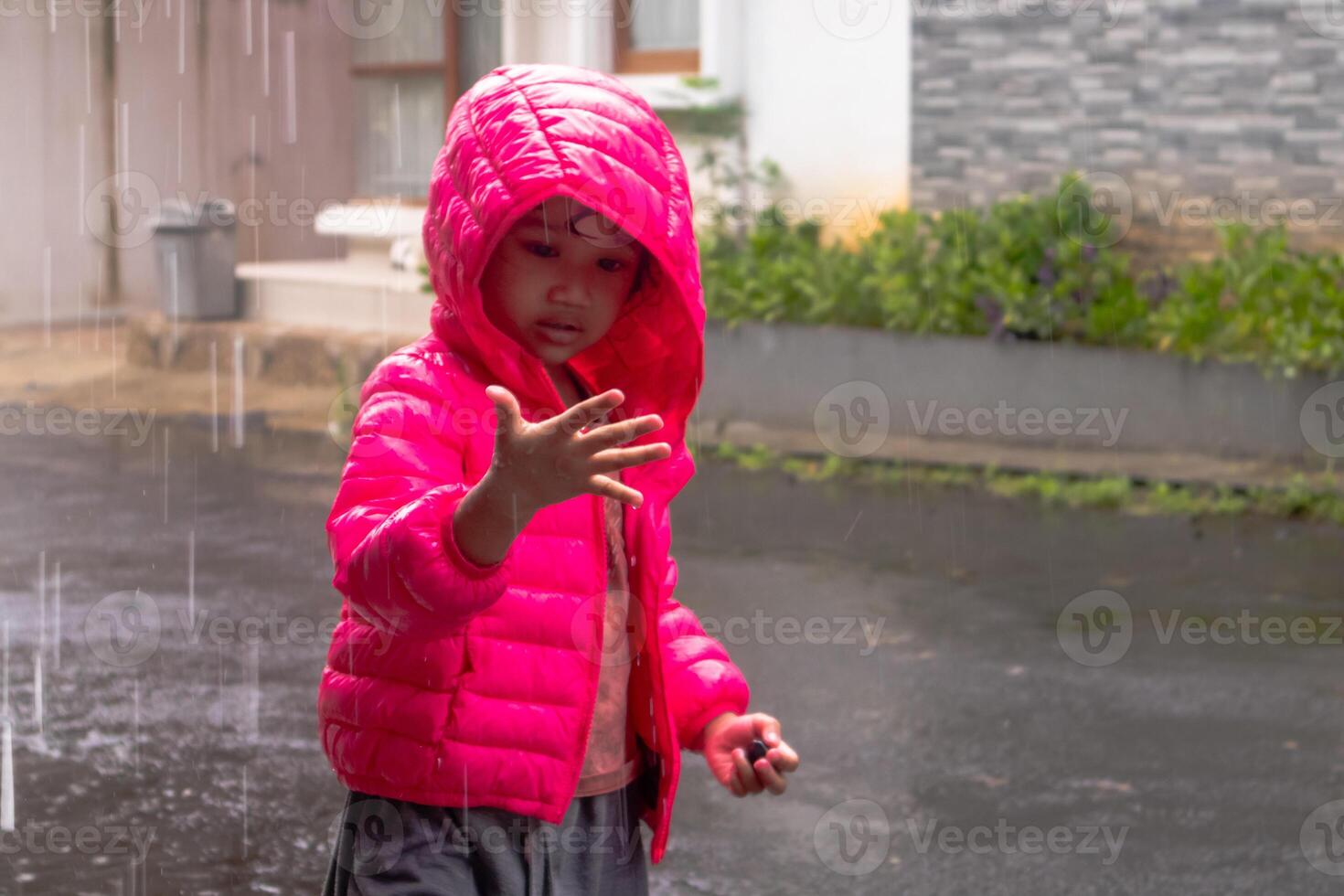 ein asiatisch wenig Mädchen tragen ein Rosa Jacke und spielen im das Regen im Vorderseite von das Haus foto