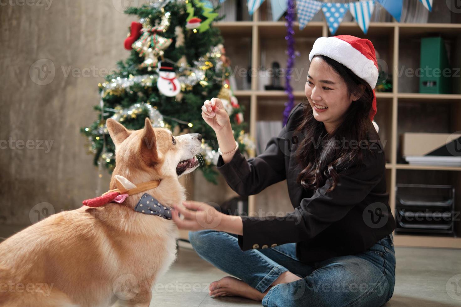 Eine junge asiatische schöne junge Arbeiterin in einem roten Hut neckt einen Hund mit Liebe auf einer Geschäftsbüroparty, dekorativ für das Feiern des Weihnachtsfestes und der Neujahrsfeiertage. foto