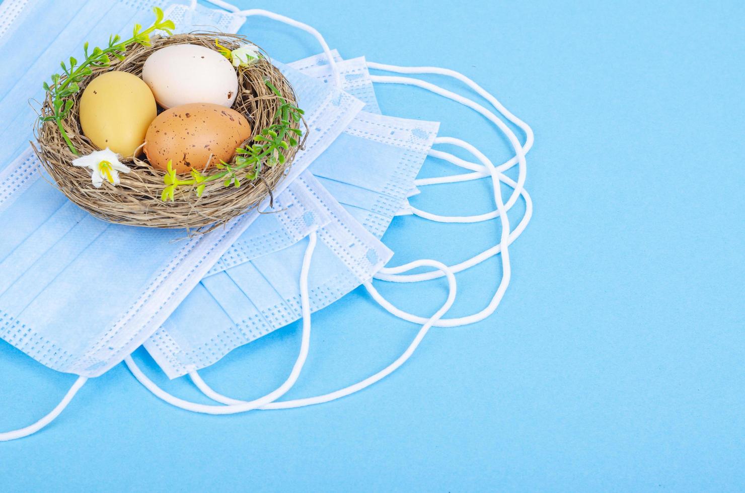 Nest mit hell gefärbten Eiern mit medizinischen Masken auf blauem Hintergrund, Platz für Text. Osterferien während der Coronavirus-Pandemie foto