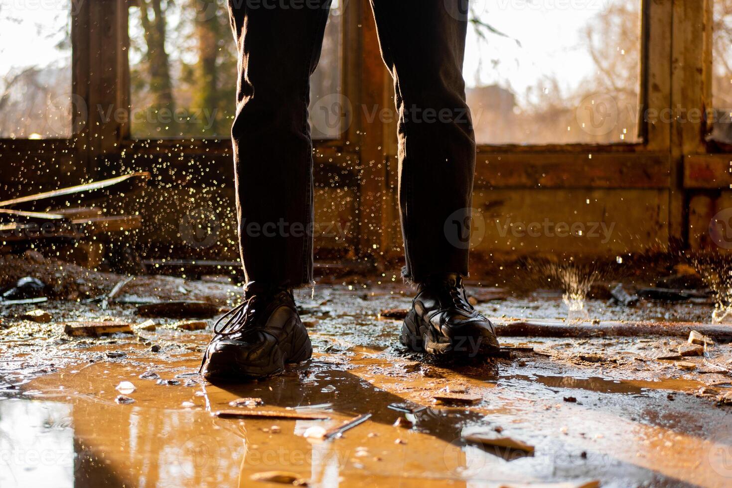 männlich Beine im schwarz Hose und Turnschuhe unter das Pfütze und gefroren Tropfen im ein überflutet, zerstört und verlassen Haus. Solar- Strahlen. Konsequenzen von ein natürlich Katastrophe. foto