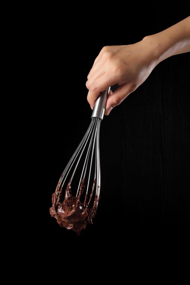 Koch hält Schneebesen und geschmolzene Schokolade auf schwarzem Hintergrund foto