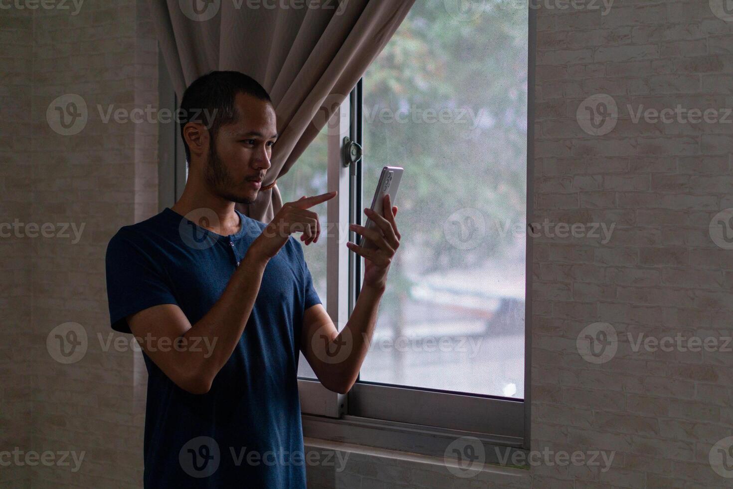 jung asiatisch Mann Stehen auf Fenster während zeigen seine Smartphone foto