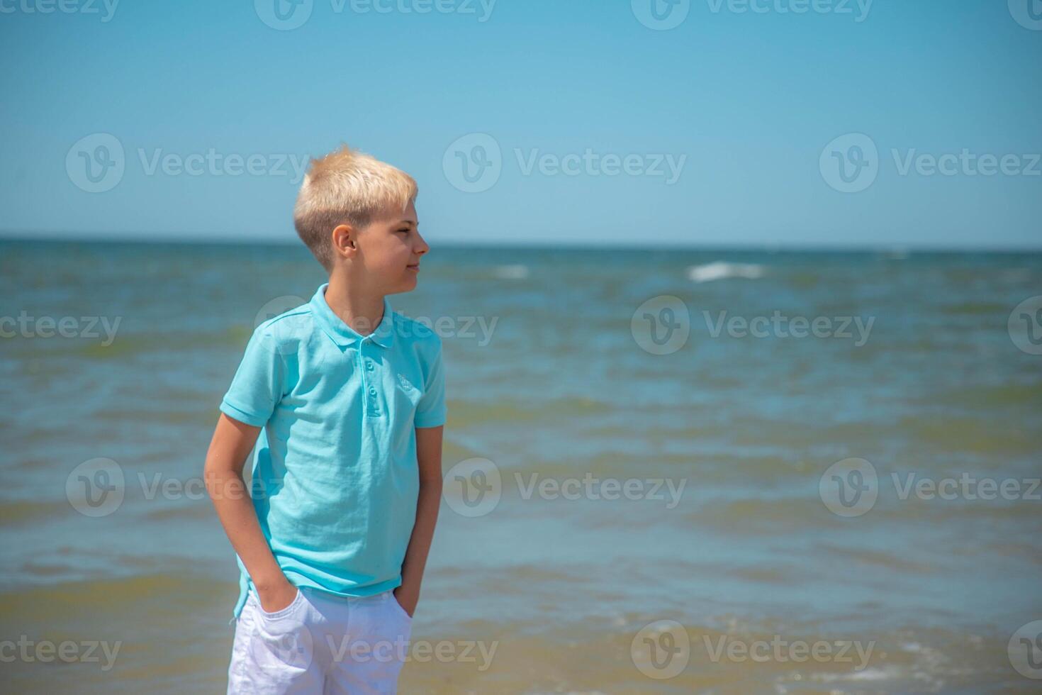 gut aussehend Teenager Junge von europäisch Aussehen mit blond Haar im ein Blau T-Shirt, und Weiß Shorts, Ständer hinter Blau Himmel, und sieht aus nachdenklich in das Distanz. Sommer- Ferien Konzept.hübsch Teenager Junge Porträt konzept.kopie Raum. foto