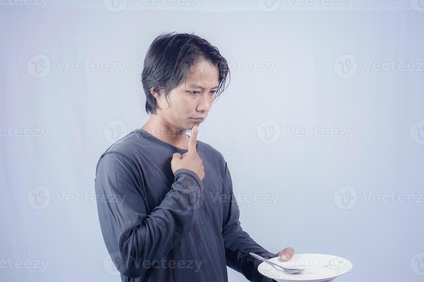 gut aussehend asiatisch Mann halten leeren Teller während Denken zum Portion Essen Speisekarte auf isoliert Weiß Hintergrund zum Werbung Speisekarte. Speisekarte Präsentation Konzept. foto