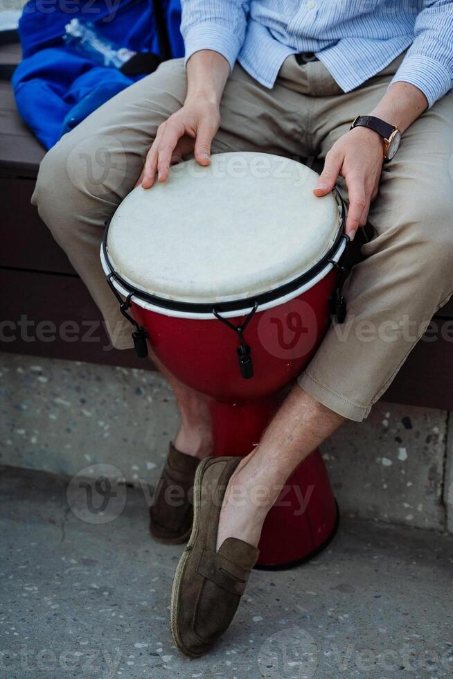 ein Büro Arbeiter gepflückt oben ein Trommel, ein Kerl im Hose und Turnschuhe Sitzung auf ein Bank spielen ein Djemba, ein afrikanisch Trommel, ein Musical Schlagzeug Instrument. foto
