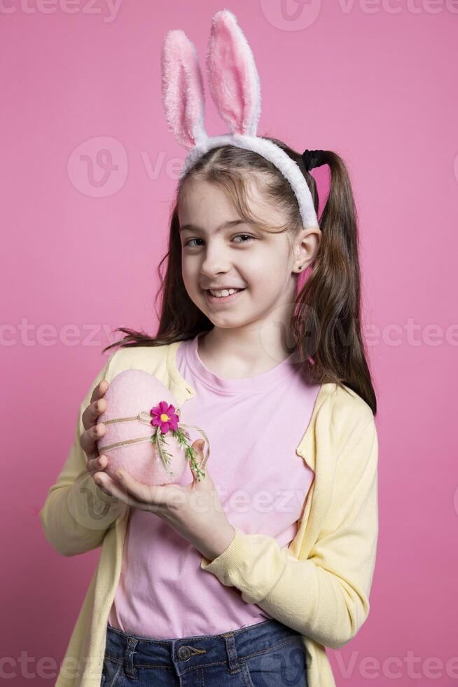 glücklich positiv Mädchen mit Hase Ohren hält ein Rosa Ostern Ei im Studio, Gefühl stolz Über ihr festlich Dekorationen handgefertigt. klein Kind Sein zuversichtlich auf Kamera, Frühling Urlaub Fall. foto