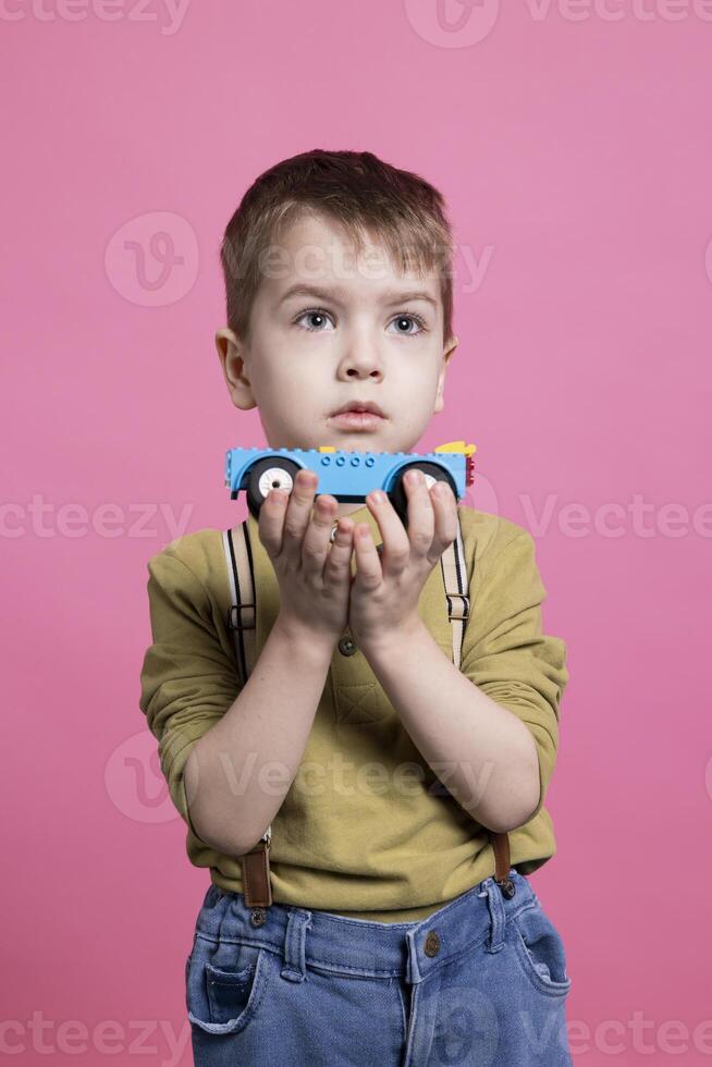 Kleinkind spielen mit ein Blau Automobil Spielzeug im Vorderseite von das Kamera, zufrieden Kind haben Spaß gegen Rosa Hintergrund. bezaubernd Kind liebend abspielen mit hell farbig Fahrzeug im Studio. foto