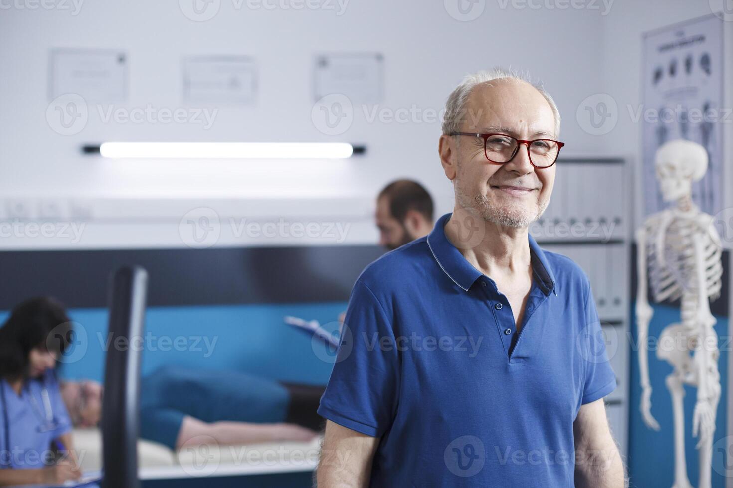 Alten Mann mit Brille Stehen im ein Physiotherapie Einrichtung. im Ruhestand Senior geduldig lächelnd beim Kamera wie er bekommt bereit zum seine Wiederherstellung beim Klinik zum Rehabilitation Behandlung durch Fitness. foto