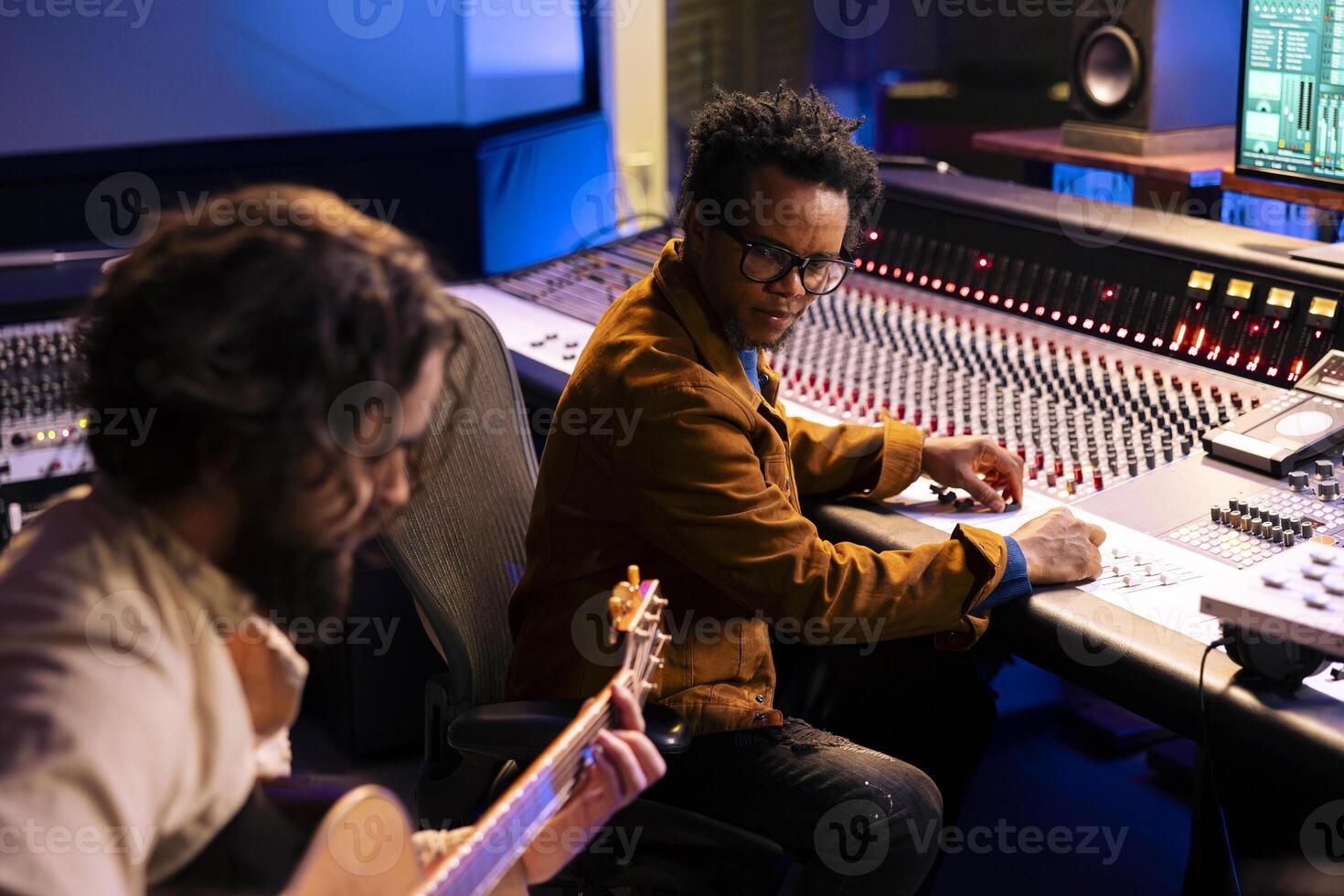 Künstler spielen ein Neu Aufzeichnung auf seine Gitarre im Steuerung Zimmer, Aufzeichnung und produzieren Musik- mit Audio- Ingenieur im Studio. Musiker Singen auf Elektro akustisch Instrument Nächster zu Mischen Konsole. foto