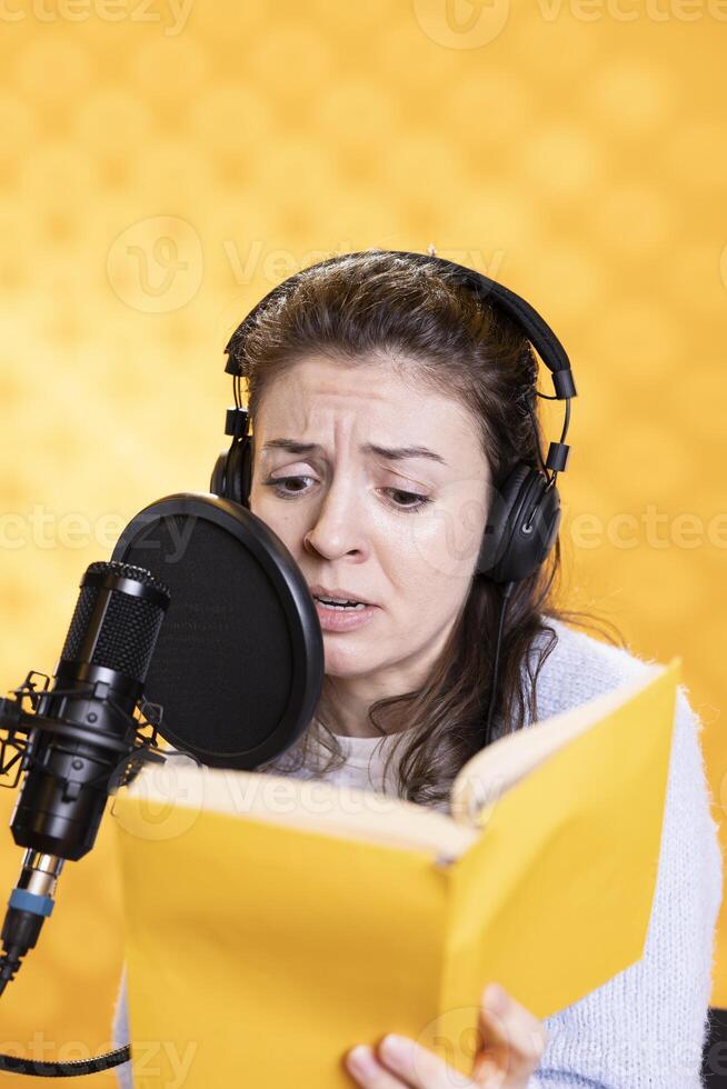 Frau mit Stirnrunzeln auf Gesicht tragen Kopfhörer lesen laut von Buch in mic gegen Hintergrund. Stimme Darsteller Aufzeichnung Hörbuch, Erstellen einnehmend Inhalt zum Zuhörer, finster zum dramatisch bewirken foto