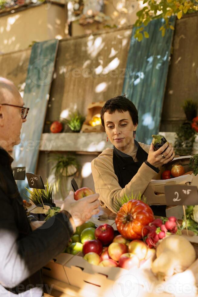 weiblich Verkäufer arrangieren organisch produzieren auf grüner Markt Stand während assistieren Alten männlich Klient mit Einkauf frisch Früchte und Gemüse. Senior Mann Kauf örtlich gewachsen bio Essen Produkte. foto