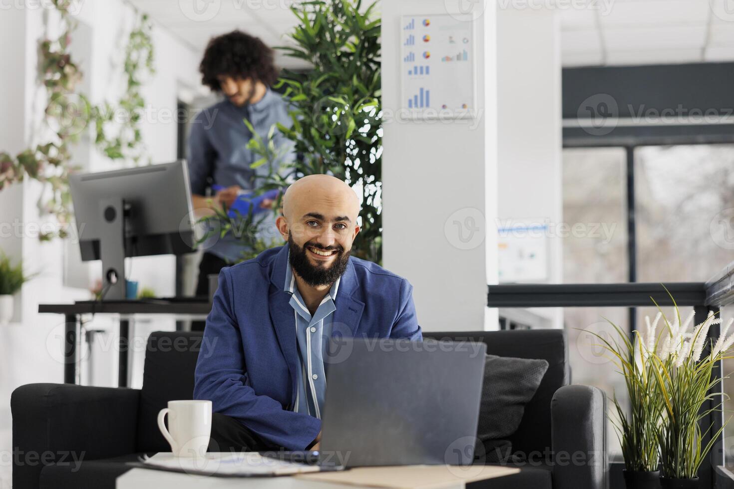 lächelnd Geschäft Unternehmer Arbeiten auf Laptop und suchen beim Kamera im Start oben Unternehmen Büro. erfolgreich Projekt Manager mit tragbar Computer während Sitzung auf Couch Porträt foto
