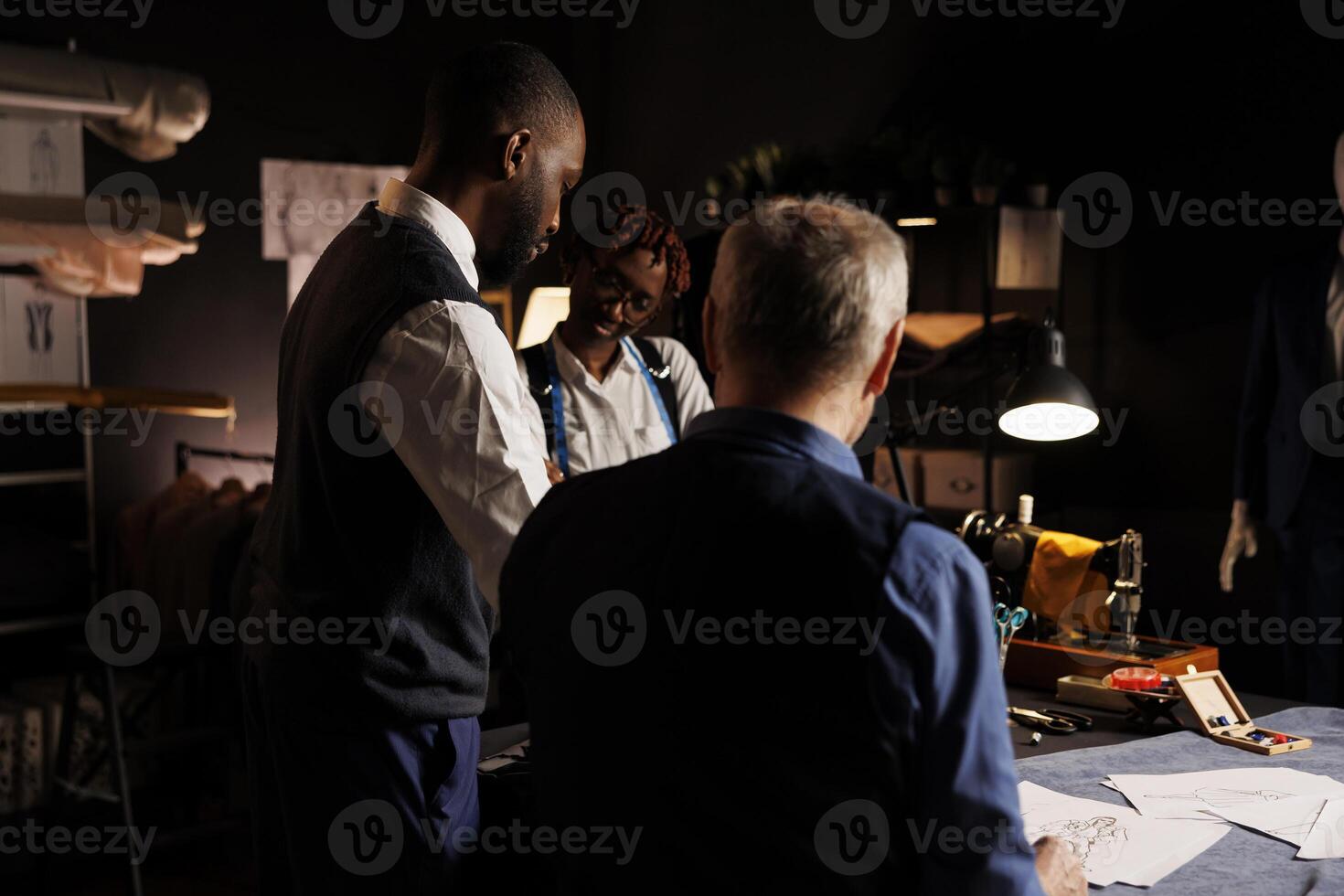 Experte Schneider Beratung mit elegant afrikanisch amerikanisch Klient auf Beste Kurs von Aktion zum seine gewünscht Hochzeit Anzug. Schneider zeigen Gentleman Skizzen zum Benutzerdefiniert gemacht maßgeschneidert Kleidung foto