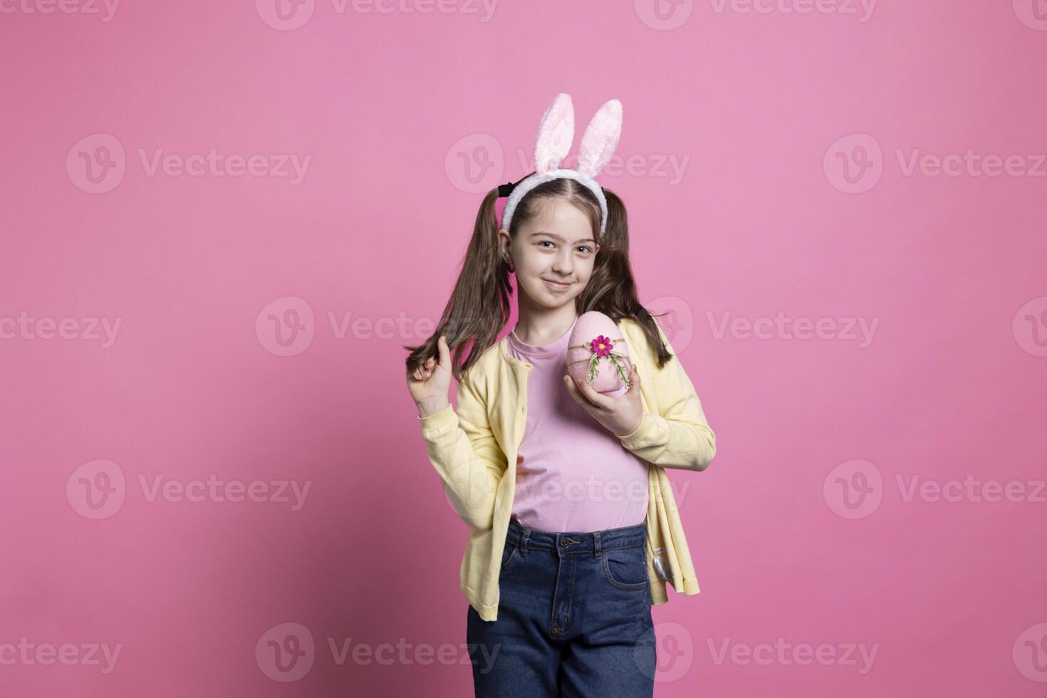 wenig jung Mädchen mit flauschige Hase Ohren zeigt an ein Rosa Ostern Ei im Vorderseite von Kamera, halten ihr handgemacht festlich Frühling Ornamente Über Studio Hintergrund. klein Kind mit Anordnungen. foto
