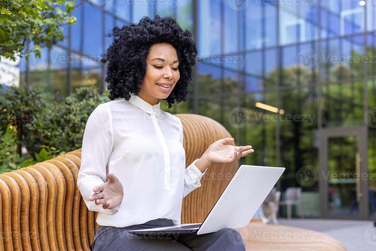 ein jung afrikanisch amerikanisch Geschäftsfrau sitzt auf ein Bank in der Nähe von ein Büro Center und Gespräche auf ein Anruf auf ein Laptop auf ihr Schoß. foto