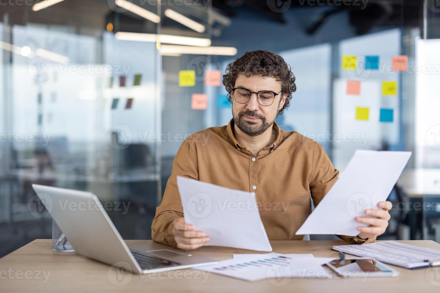 ein konzentriert männlich Geschäft Fachmann Bewertungen wichtig Papiere während Arbeiten beim ein Schreibtisch im ein zeitgenössisch Büro Einstellung. das Arbeitsplatz ist ausgestattet mit ein Laptop und sichtbar klebrig Anmerkungen. foto