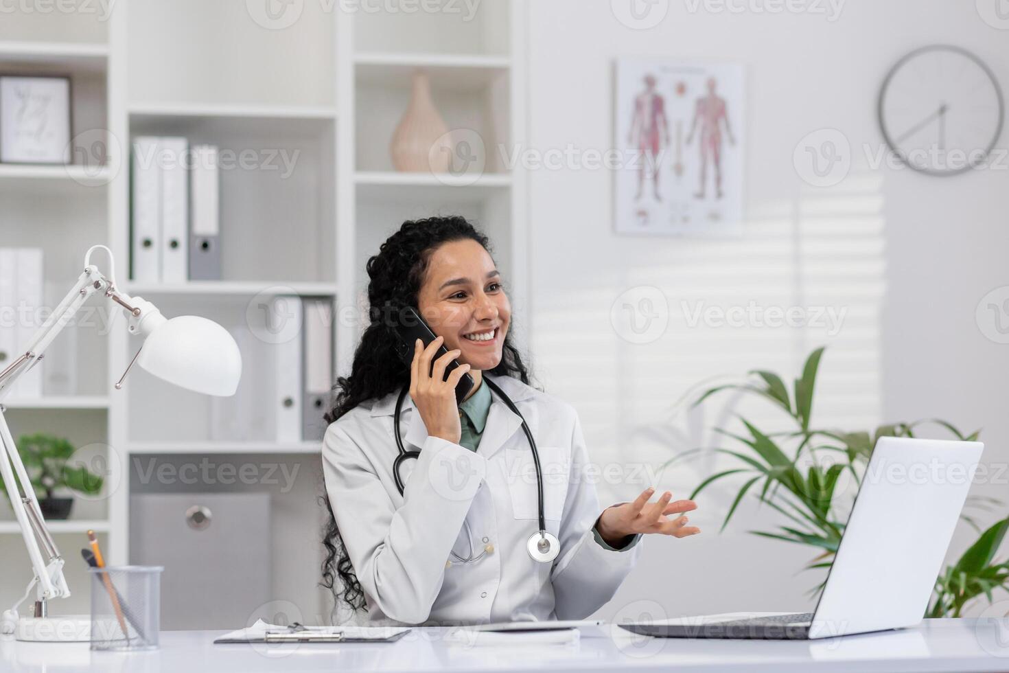 ein heiter weiblich Arzt mit lockig Haar glücklich reden auf das Telefon während suchen beim ihr Laptop im ein gut beleuchtet, organisiert Büro. foto