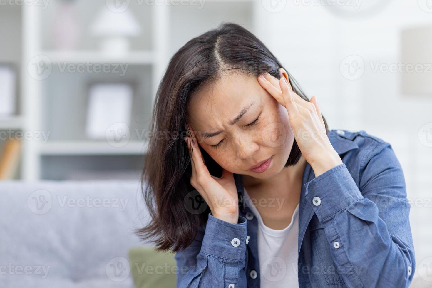 krank Frau allein beim heim, asiatisch deprimiert Frau haben stark Kopfschmerzen, Sitzung auf Sofa im Leben Zimmer beim heim, halten Hände zu Kopf von Schmerz. foto