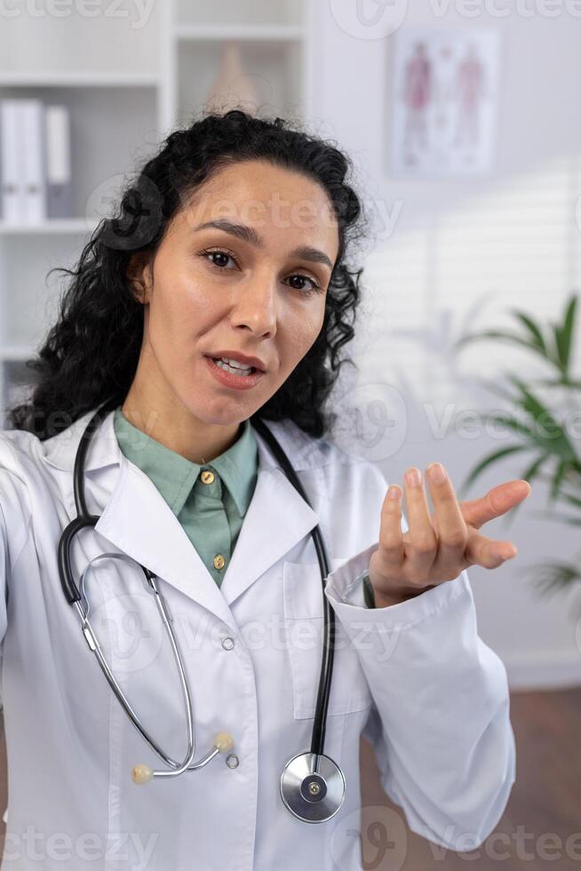 Vertikale Schuss, weiblich Arzt Ernsthaft reden und Beratung geduldig suchen entfernt in Telefon Kamera, mit Smartphone Anruf Anwendung, Arbeiten Innerhalb Klinik Büro. foto