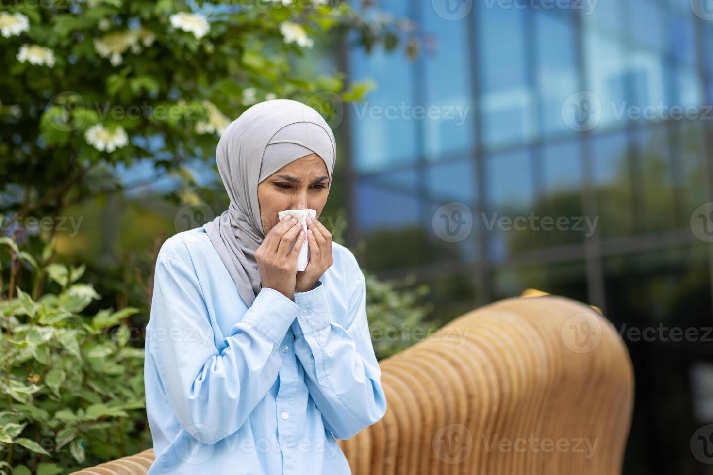 ein jung Frau im ein Blau Hijab Gefühl nicht wohl, Niesen in ein Gewebe im ein städtisch Park Rahmen mit Grün. foto