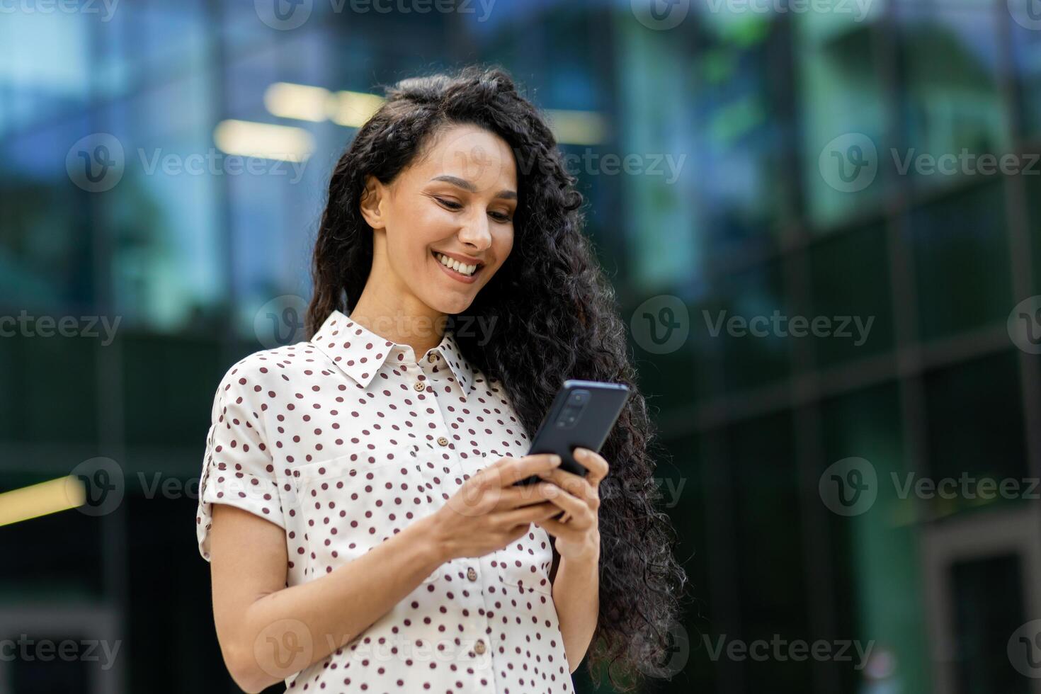 jung, attraktiv spanisch Geschäftsfrau mit lockig Haar prüft ihr Telefon mit ein Lächeln während nehmen ein gemächlich gehen während ihr Mittagessen Unterbrechung. foto