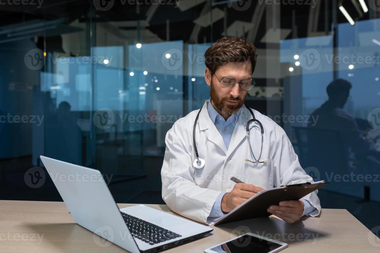 ein jung männlich Arzt im ein Mantel, tragen Brille und ein Stethoskop funktioniert im ein Büro im ein Krankenhaus beim ein Tabelle mit ein Laptop. schreibt das Geschichte von Selten Dinge, funktioniert mit Unterlagen, hält ein Ordner. foto