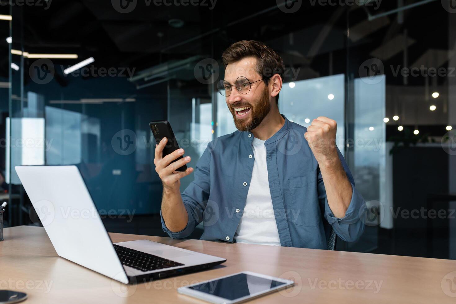 erfolgreich reifen Geschäftsmann Boss Arbeiten Innerhalb modern Büro Gebäude beim Schreibtisch, mit Laptop, Mann mit Bart und Brille halten Smartphone, habe gut Nachrichten Über Sieg Triumph feiern. foto