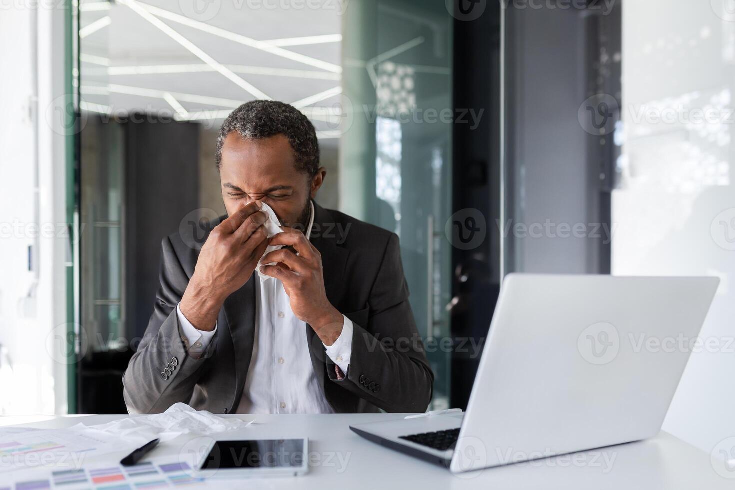 krank Mann beim Arbeitsplatz Innerhalb Büro mit Freude und kalt, Geschäftsmann Niesen Sitzung beim Schreibtisch, afrikanisch amerikanisch Mann mit kalt Verwendet Laptop beim arbeiten. foto