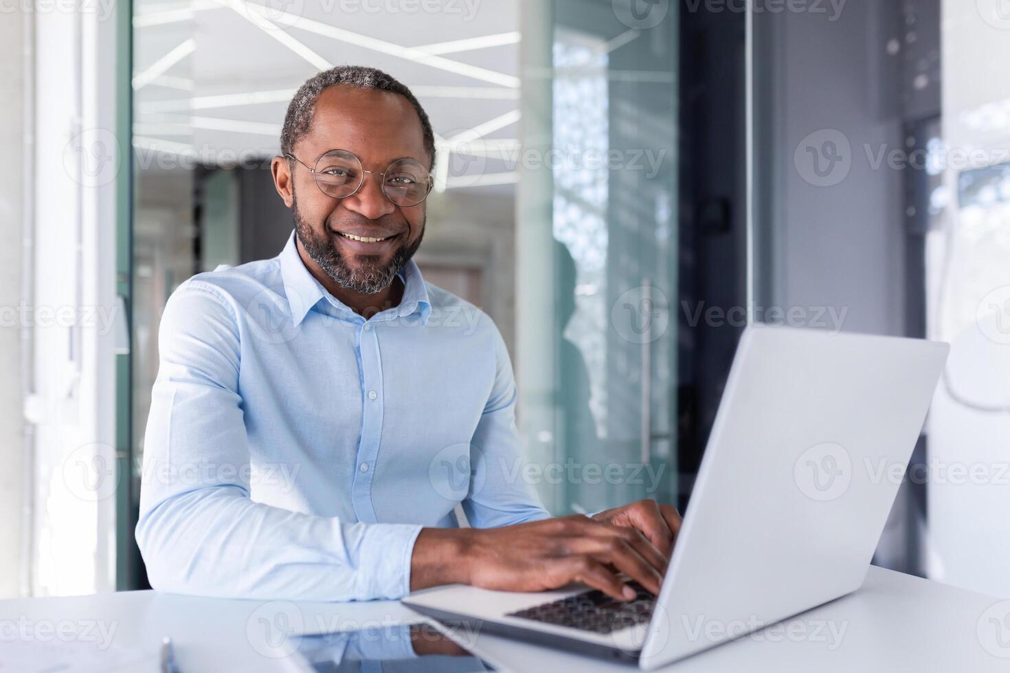 erfolgreich afrikanisch amerikanisch Geschäftsmann im Hemd und Brille Arbeiten Innerhalb hell modern Büro, Mann lächelnd und suchen beim Kamera, Porträt von erfolgreich Boss mit Laptop Sitzung beim Arbeitsplatz. foto