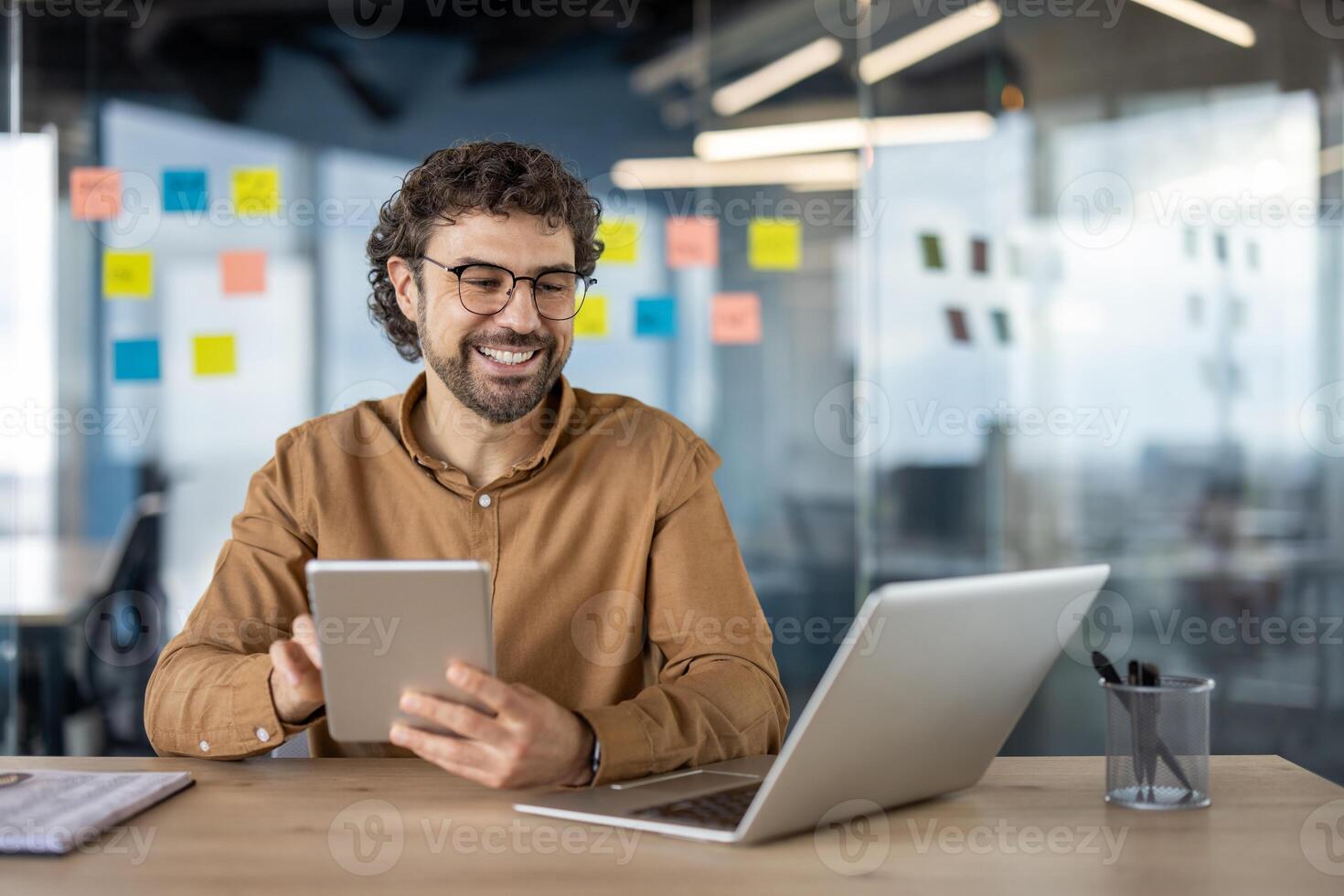 lächelnd männlich Fachmann Multitasking mit ein Tablette und Laptop im ein modern Büro Rahmen mit klebrig Anmerkungen im das Hintergrund. foto