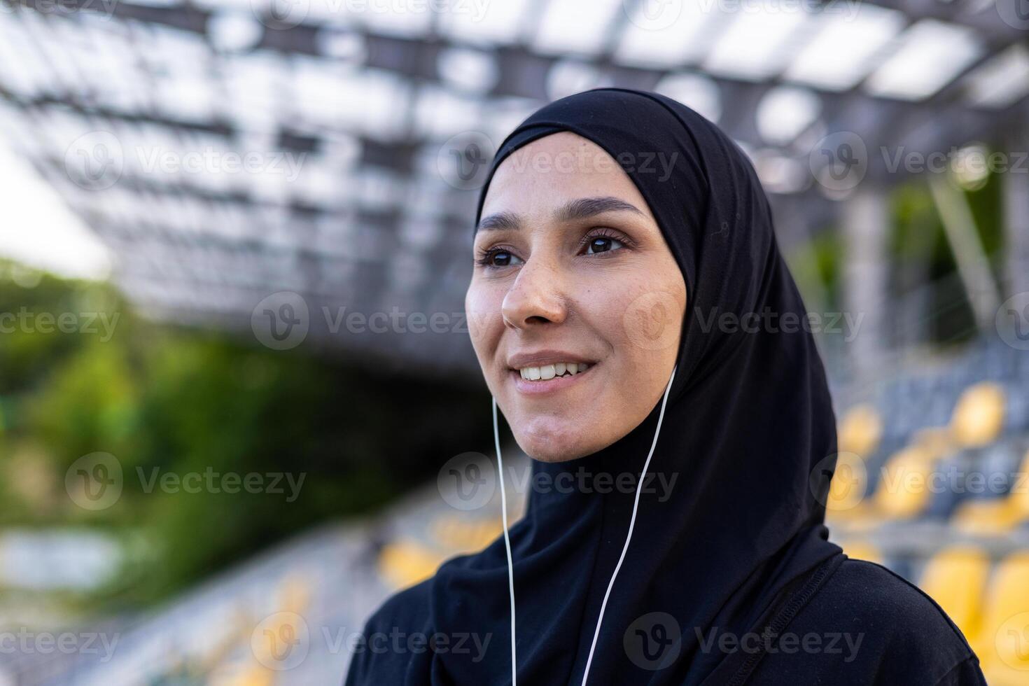 Porträt von Muslim Frau im schwarz Kleider und Hijab Gefühl inspiriert während Hören zu Musik- im verdrahtet Kopfhörer. aktiv weiblich tun Täglich Sport Routine und suchen voraus auf verschwommen Hintergrund. foto