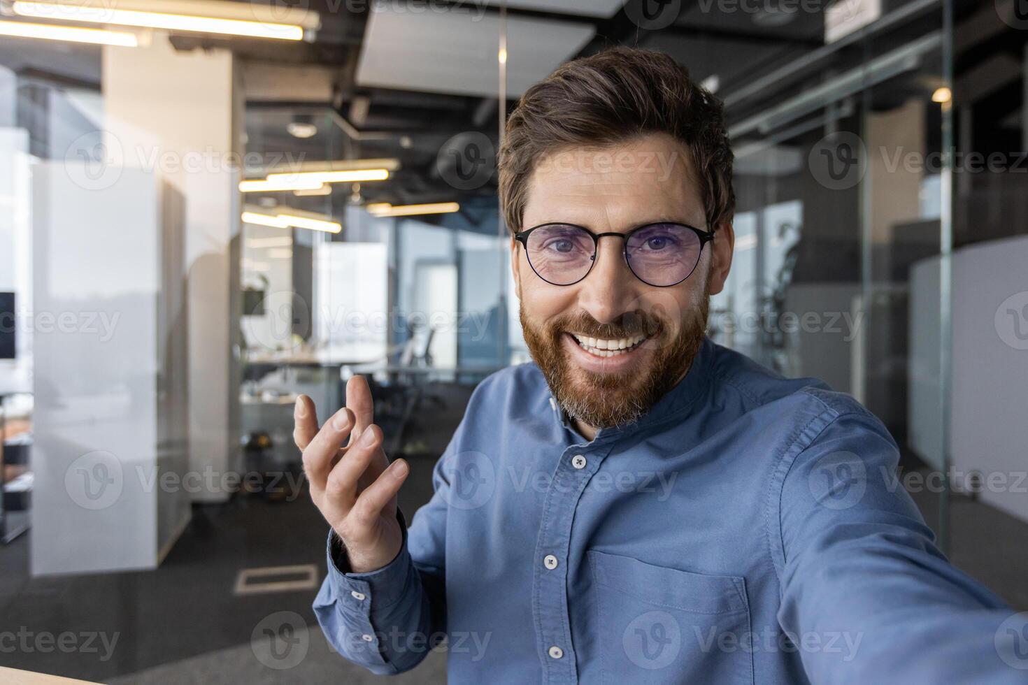 Nahansicht Foto von ein jung lächelnd Mann im das Büro reden auf ein Anruf zu das Kamera Telefon, erklären durch gestikulieren mit seine Hand.