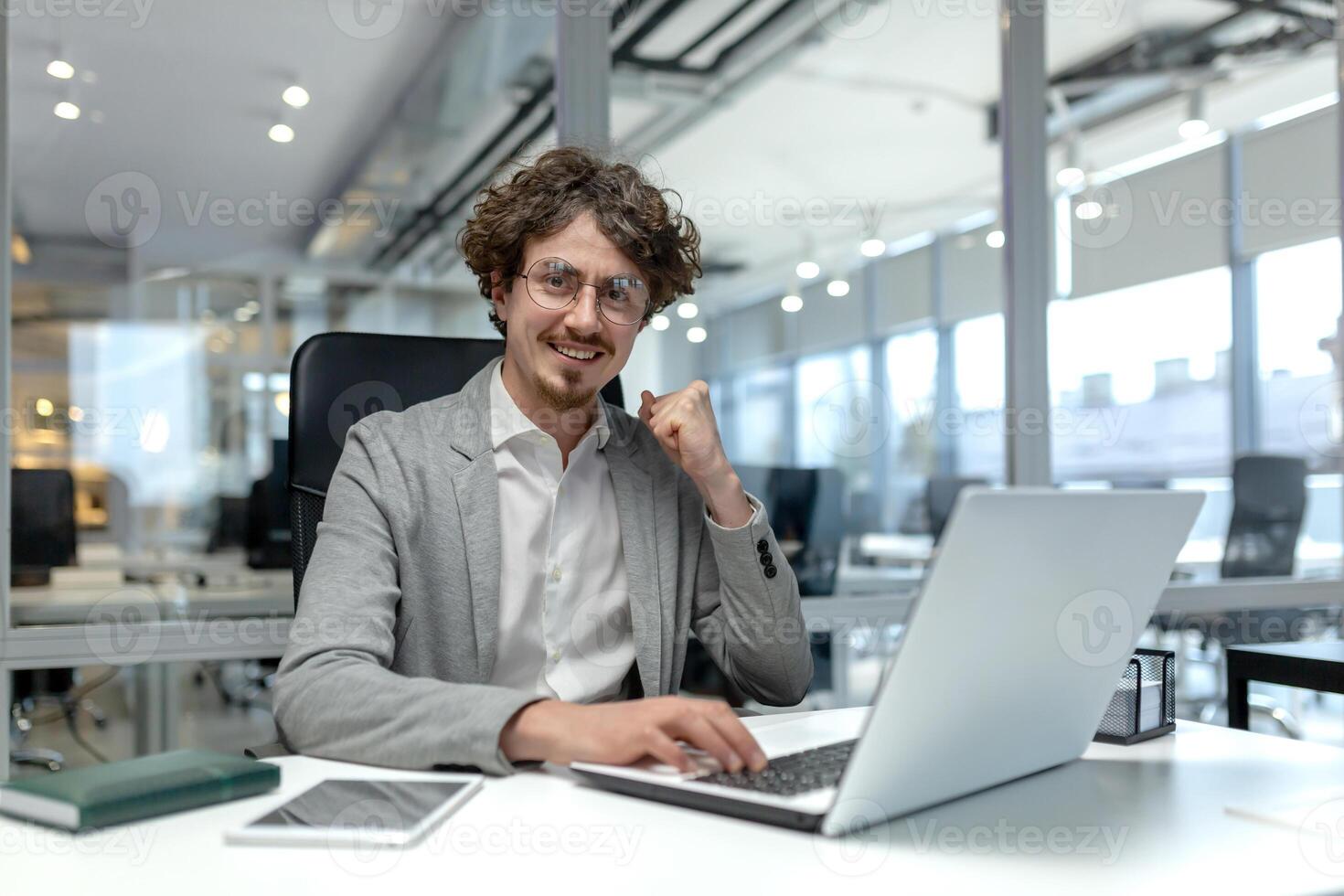lächelnd jung Geschäftsmann mit lockig Haar konzentriert auf Arbeit beim seine Laptop im ein gut beleuchtet modern korporativ Büro Einstellung. foto