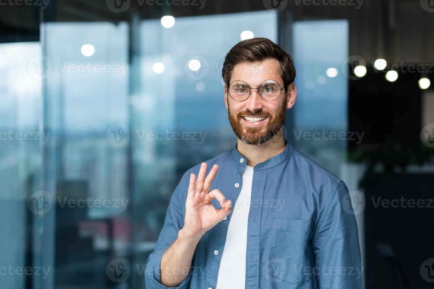 Porträt von ein erfolgreich reifen Geschäftsmann Innerhalb das Büro, ein Mann mit ein Bart und Brille im ein Blau Hemd ist lächelnd und suchen beim das Kamera, zeigen das in Ordnung Geste mit seine Hand. foto