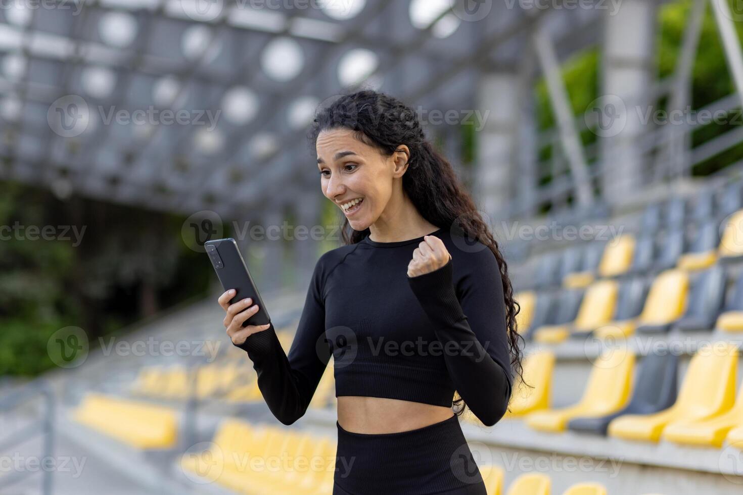 ein froh jung weiblich im sportlich tragen Prost mit ihr Telefon im Hand beim ein leeren Stadion, ausdrücken Sieg. foto